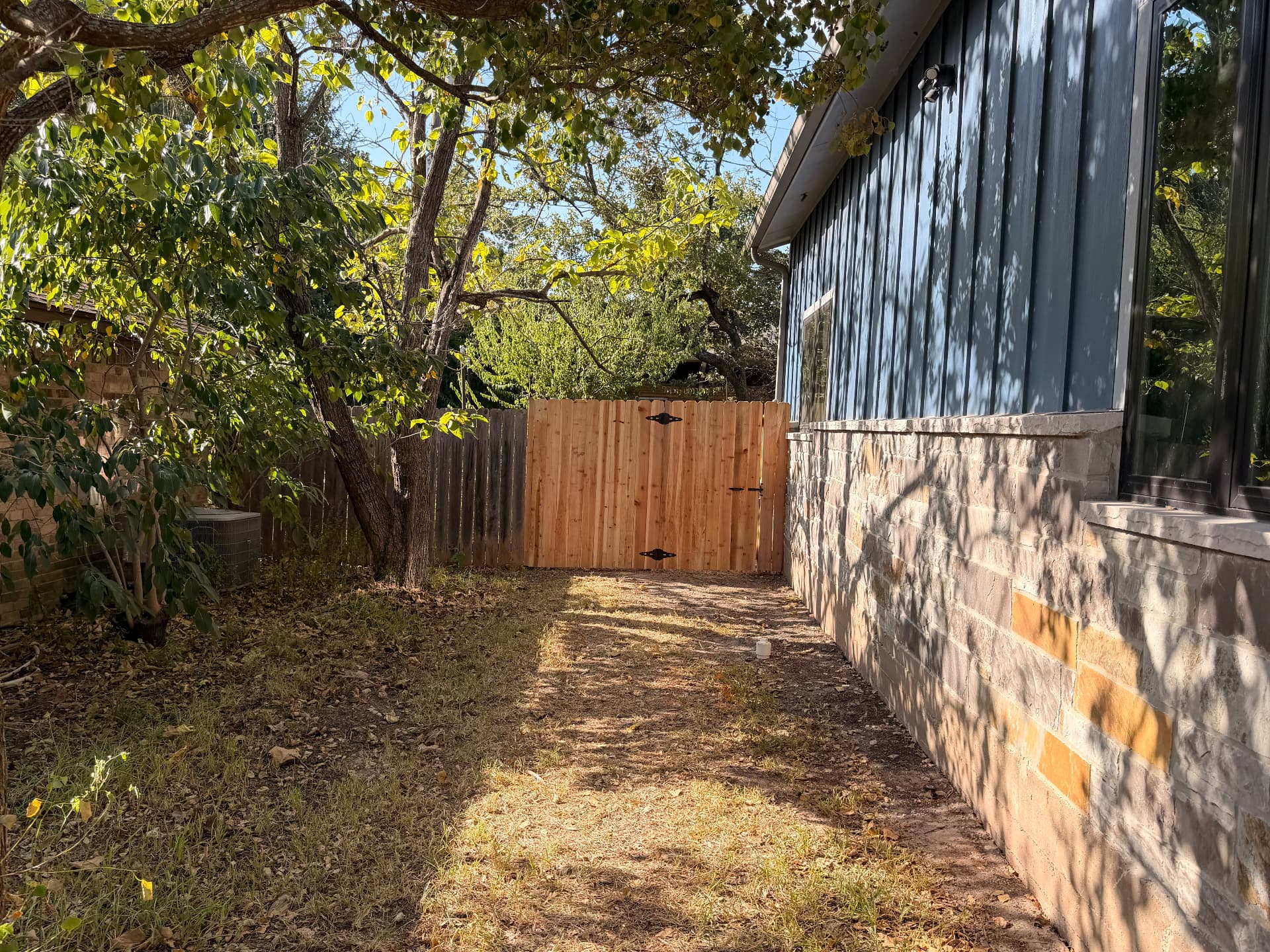 Cedar privacy fence gate beside a navy board-and-batten home with stone wainscot in Bryan TX