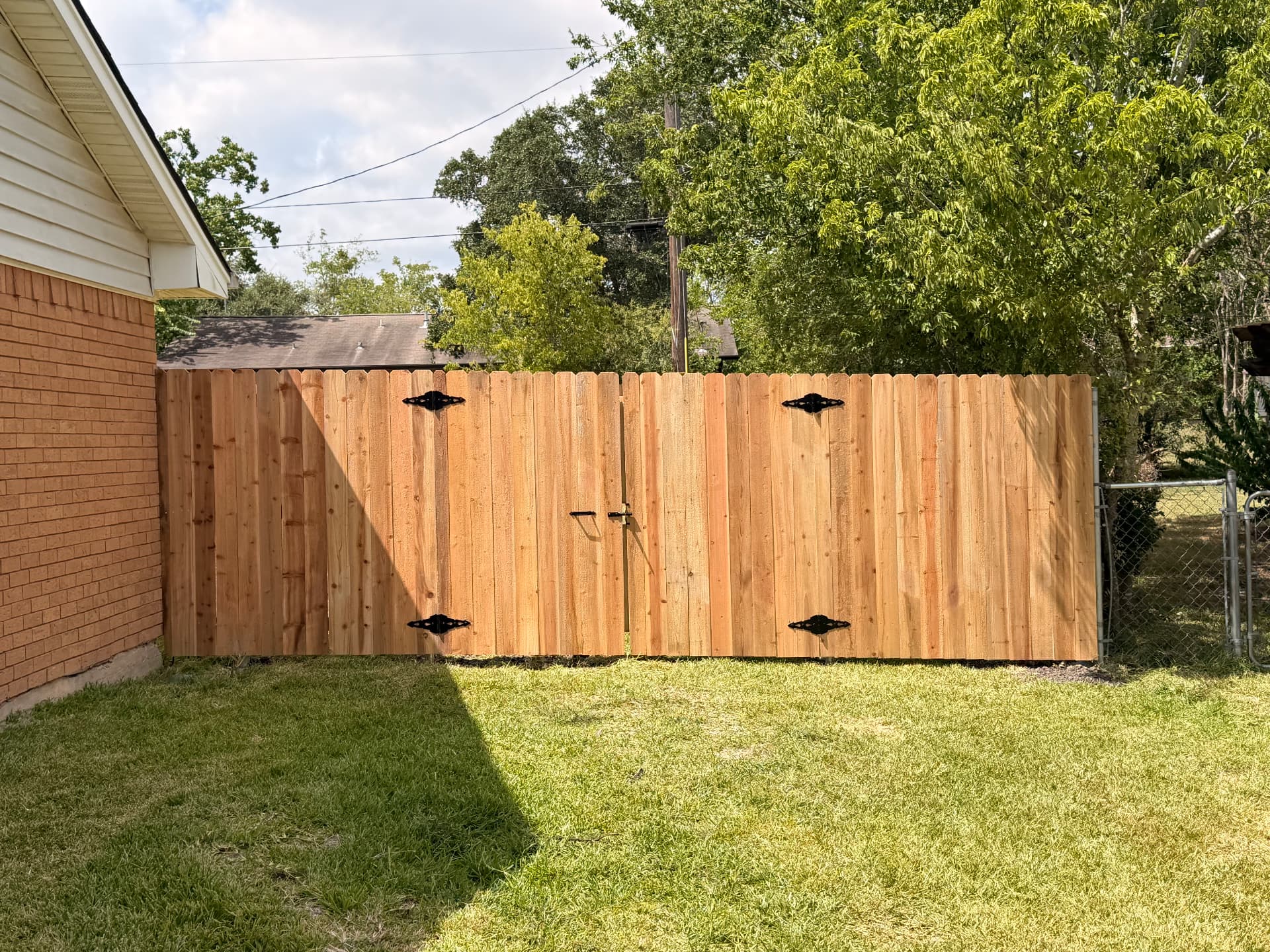 Wide cedar double drive gate with decorative black hinges beside a brick home in Bryan TX